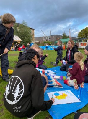 Young people gather around a tarpaulin under grey skies, working on placards with environmental slogans