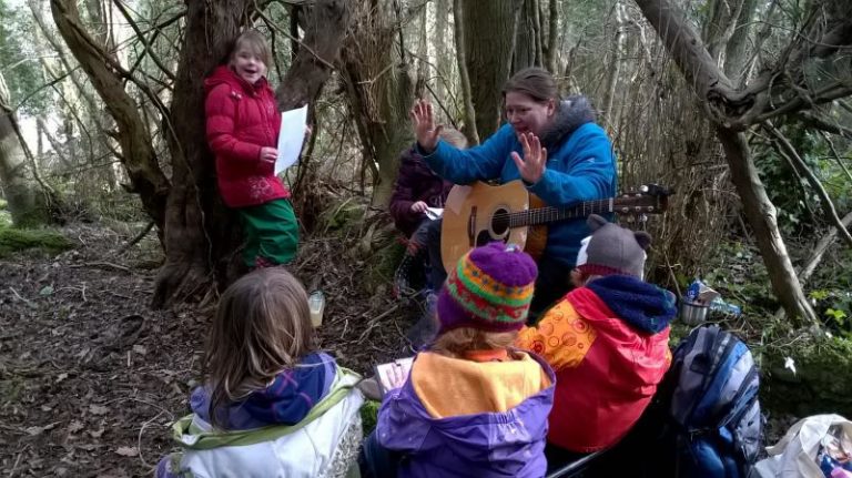 5 children surrounding an adult with a guitar in the woods